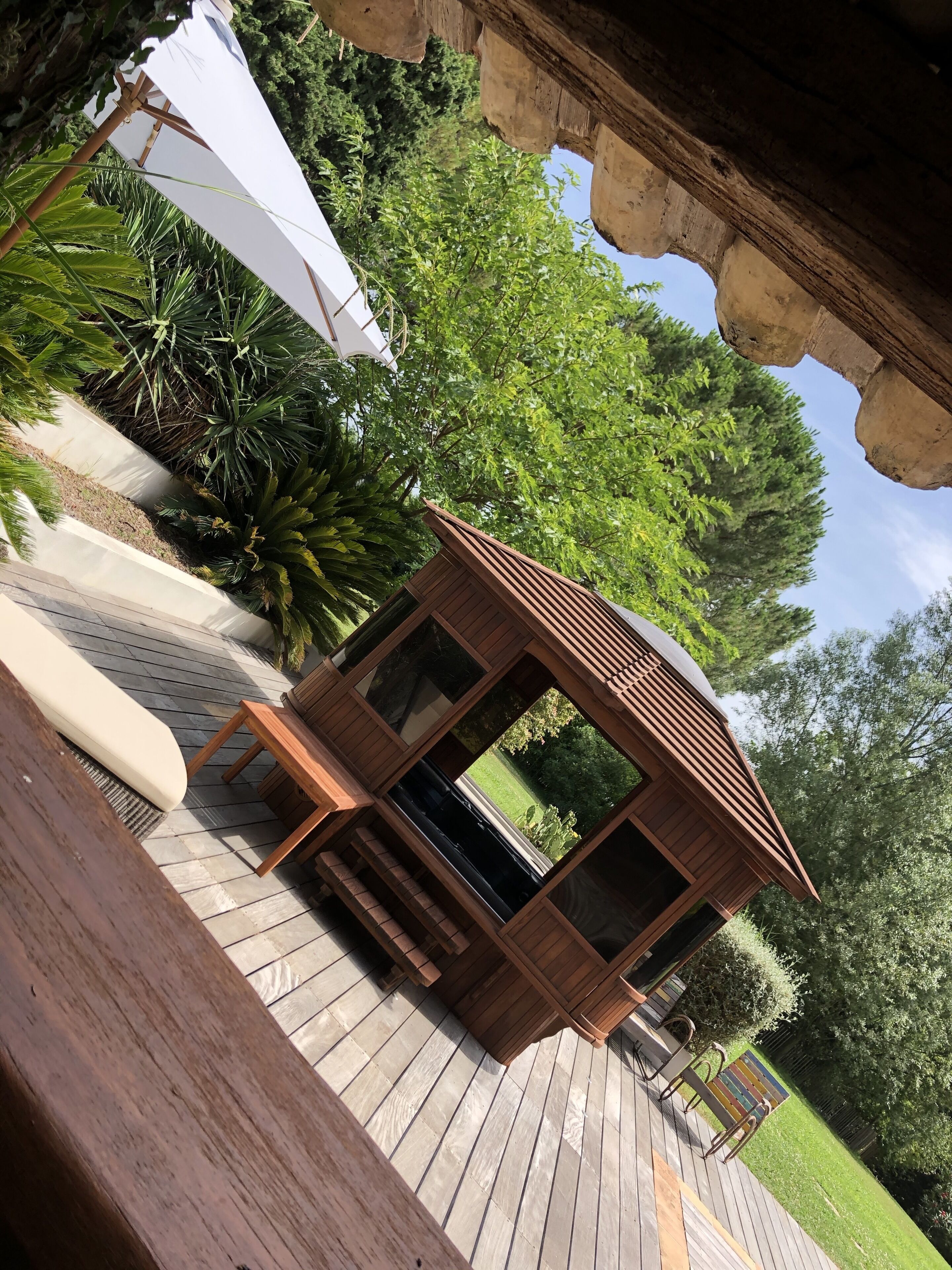 Un gazebo in legno con panchina e ombrellone, immerso nel verde e con un pavimento in legno.