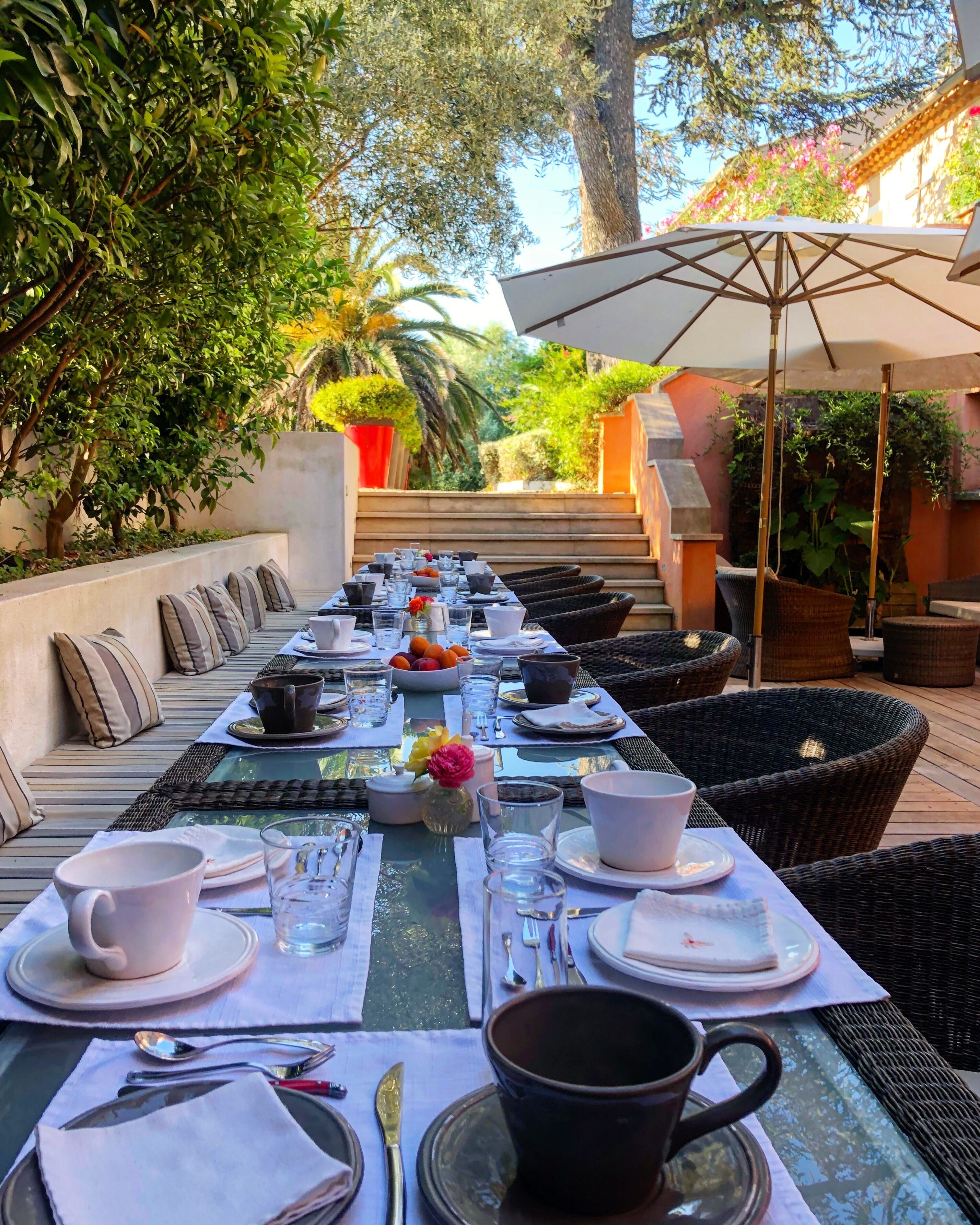 Un coin repas avec une longue table dressée pour un repas, entouré de chaises en osier et d'un grand parasol.