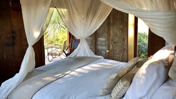 Una cama con sábanas blancas y dosel, paredes de madera y vista a muebles de exterior.