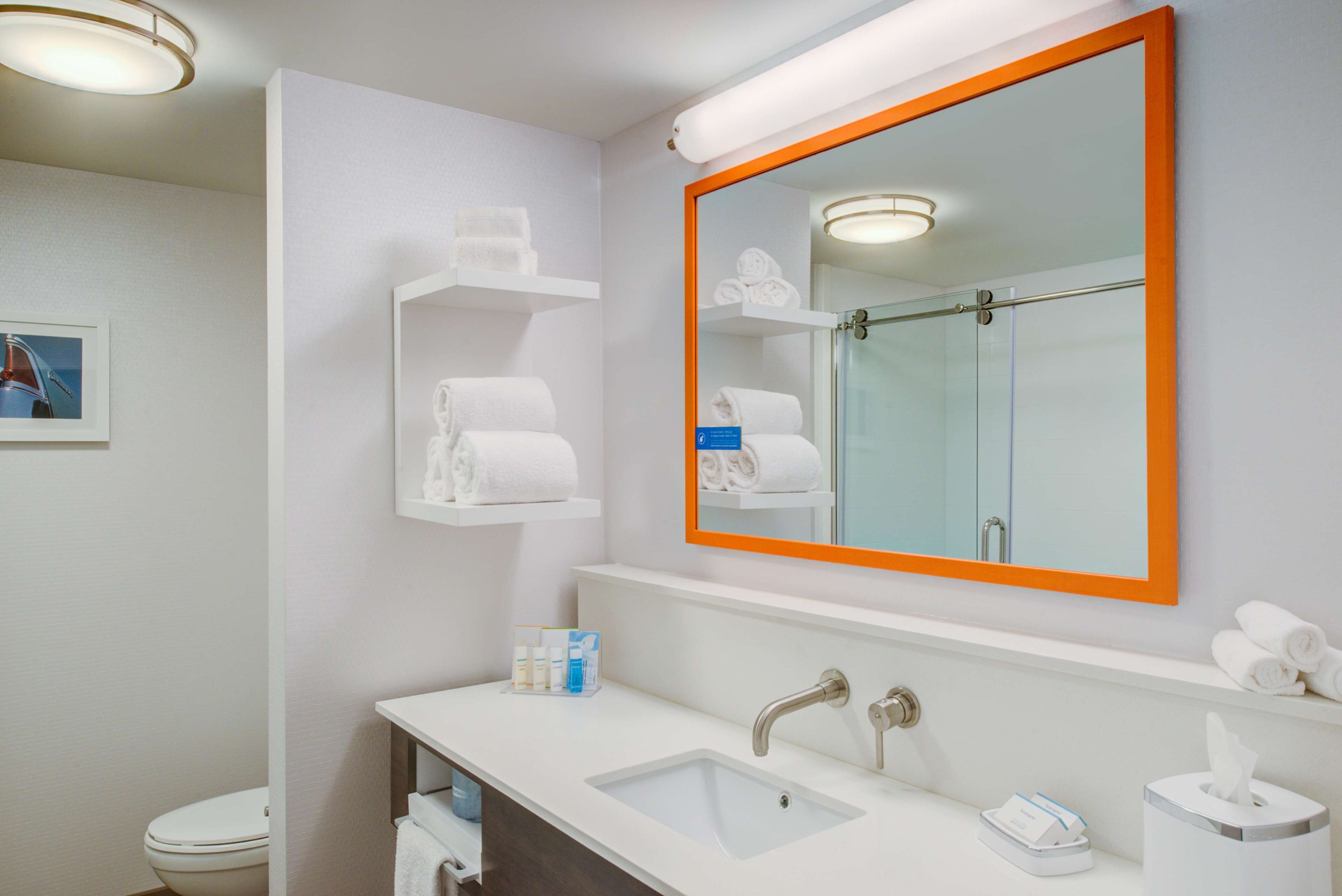 Un baño moderno con un lavamanos blanco, dos grifos, un espejo de marco naranja y estantes con toallas.