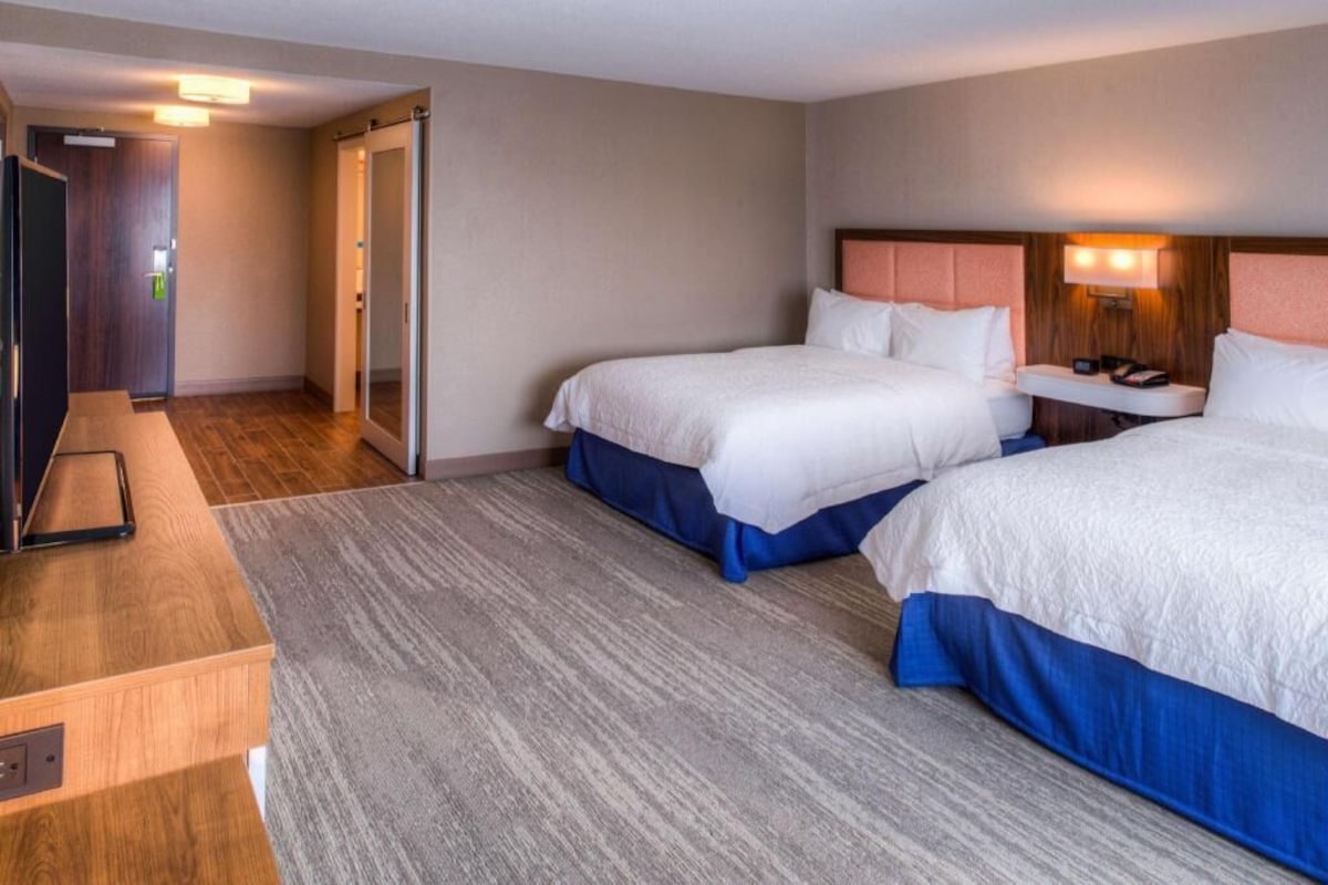 A hotel room with two beds, a desk, a chair, a nightstand, and a sliding door.