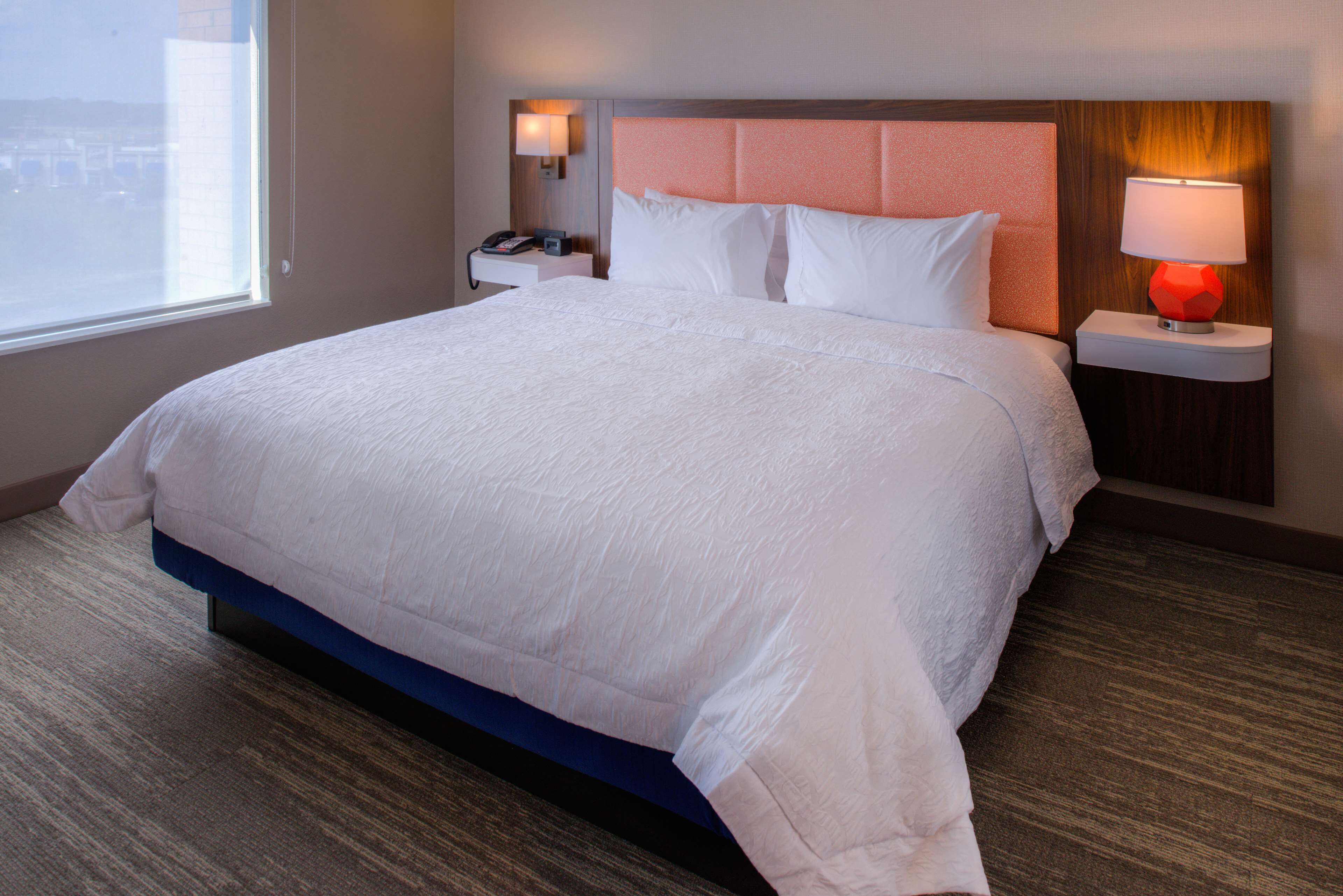 A hotel room with a large bed, a nightstand with a lamp, and a view of the city through the window.
