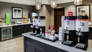 A coffee station with multiple beverage dispensers, a refrigerated display case, and a fruit basket.
