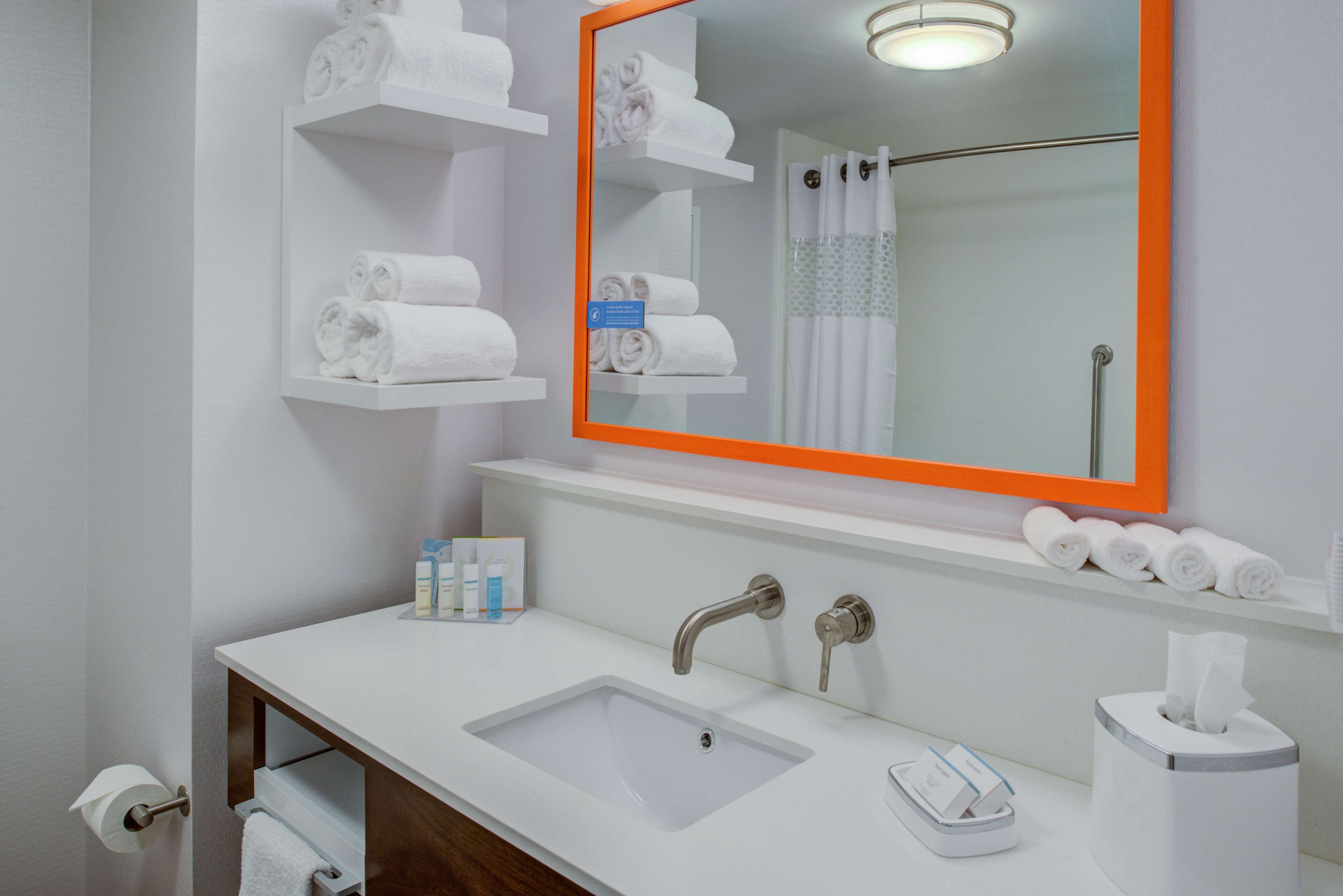 Ein Badezimmer mit einem weißen Waschbecken, zwei Wasserhähnen, einem Spiegel mit orangefarbenem Rahmen und Regalen mit aufgerollten Handtüchern.