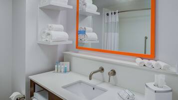 Ein Badezimmer mit einem weißen Waschbecken, zwei Wasserhähnen, einem Spiegel mit orangefarbenem Rahmen und Regalen mit aufgerollten Handtüchern.