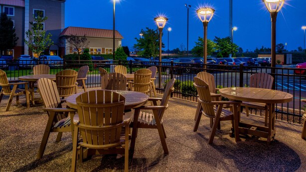 An outdoor patio area with wooden tables and chairs, surrounded by a metal fence, and illuminated by street lamps. - Hampton Inn & Suites Wixom (Wixom)