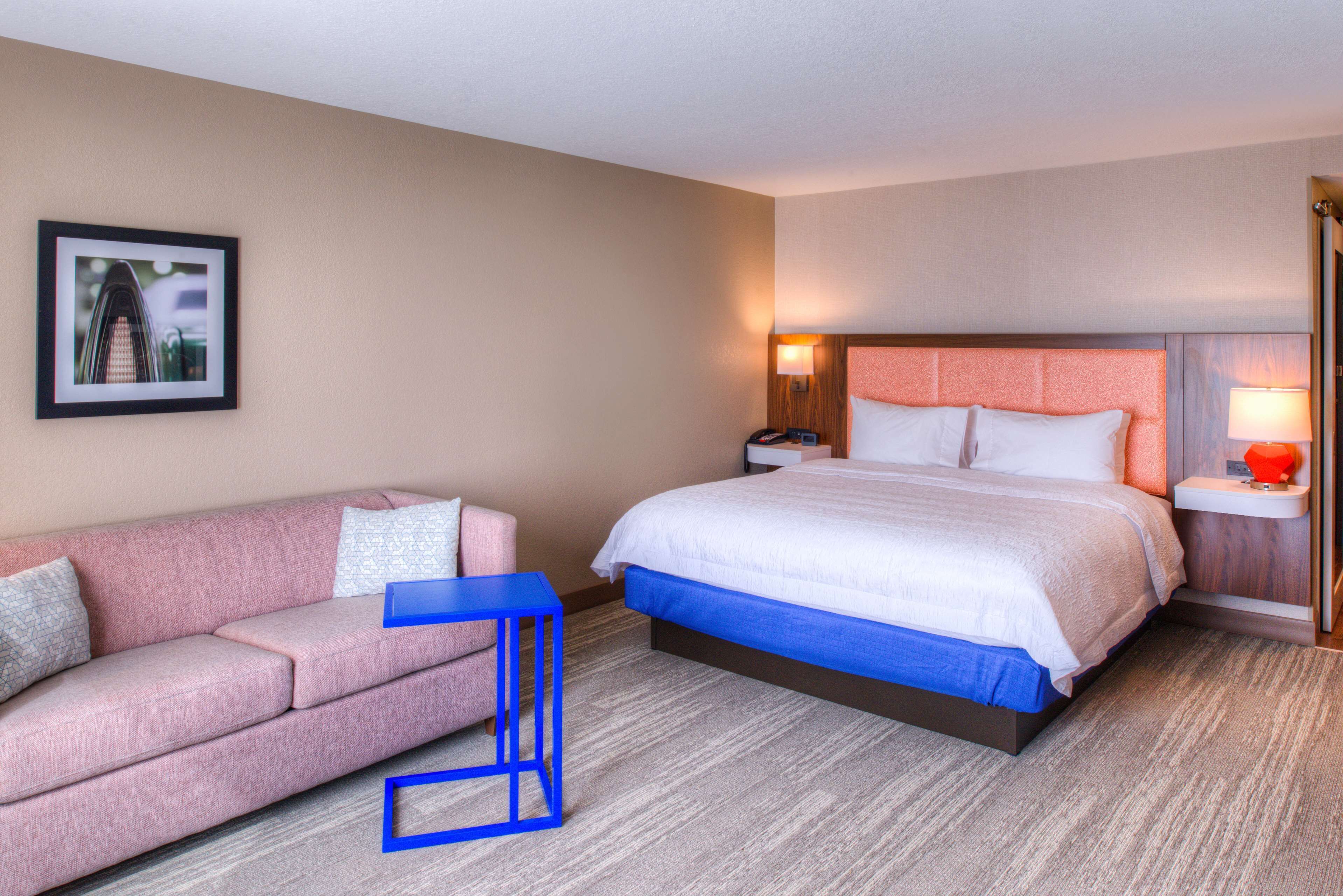 Quarto de hotel com uma cama, um sofá rosa, uma mesa azul e um quadro na parede.