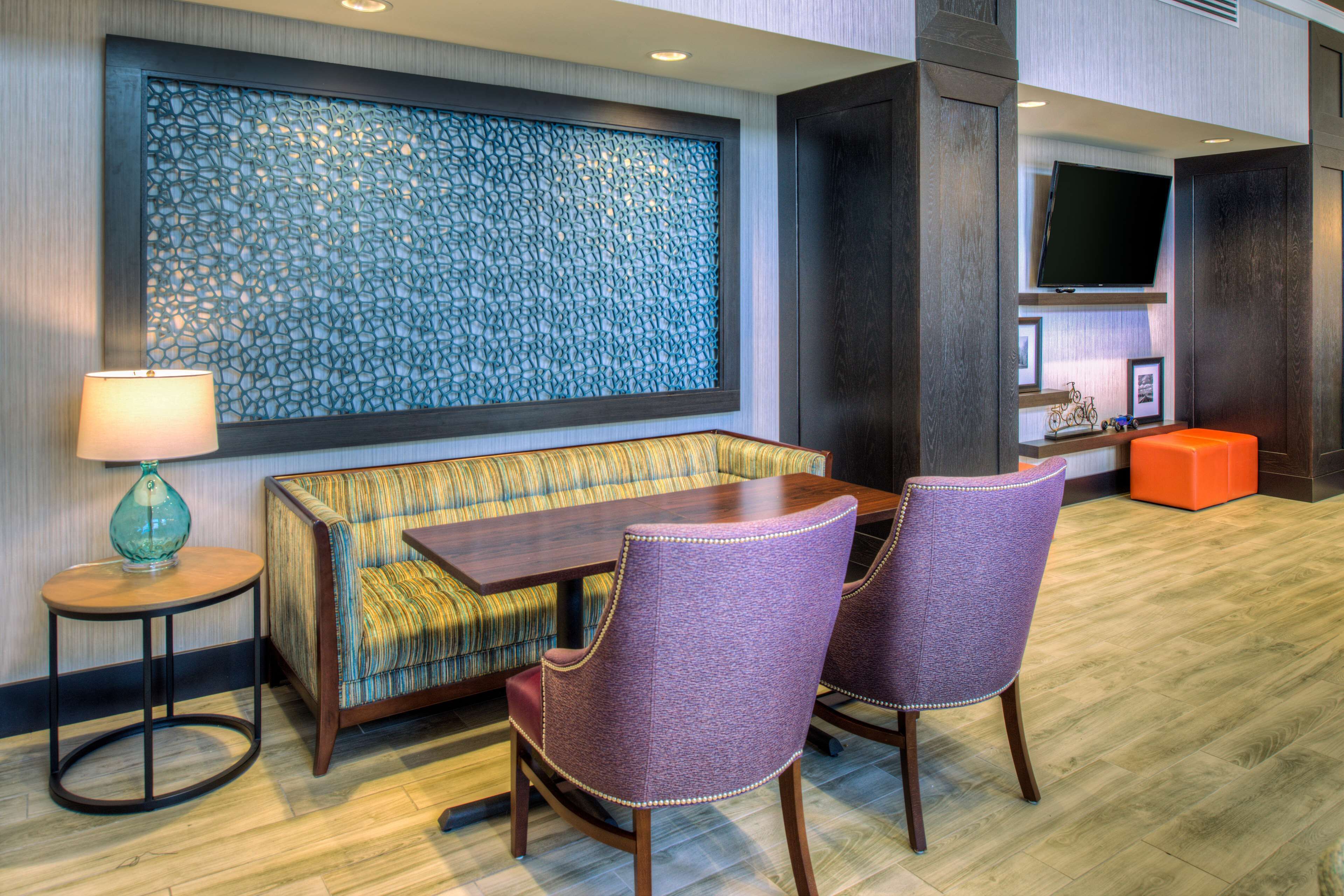 A modern hotel lobby with a sofa, two chairs, a table, a lamp, a decorative wall, and a television.
