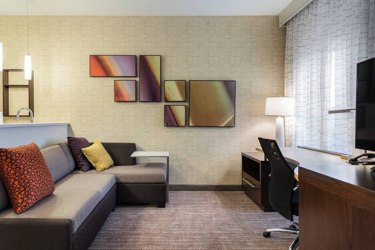 A modern hotel room with a sofa, a desk, a chair, and artwork on the wall.