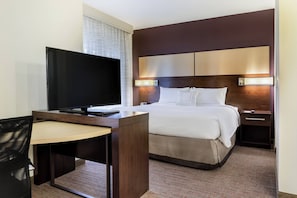 A hotel room with a bed, a desk with a TV, a chair, and a bedside table.