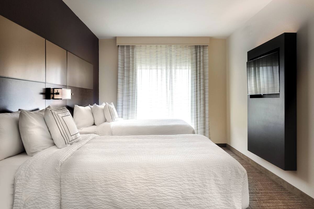 A hotel room with two beds, a large window with curtains, a flat-screen TV mounted on the wall, and a modern, minimalist design.