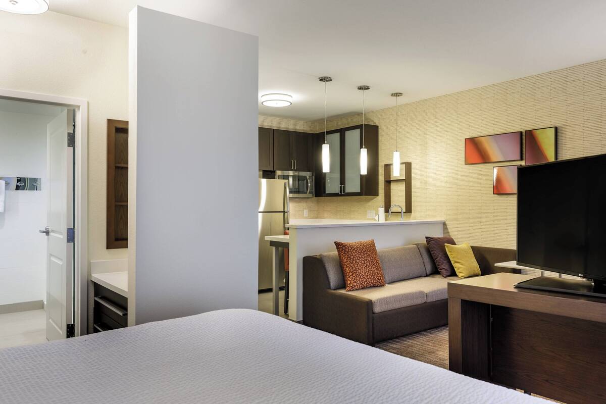 A modern hotel room with a bed, a sofa, a TV, and a kitchenette.