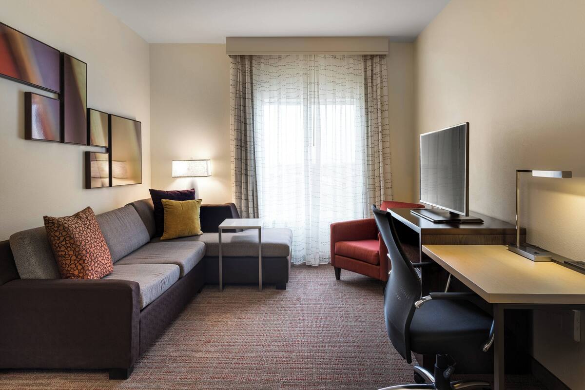 A modern hotel room with a sofa, a desk, a chair, a TV, and artwork on the wall.