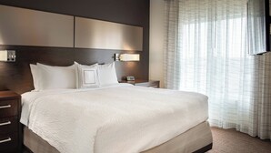 A neatly made bed with a white comforter and pillows, a wooden headboard, a bedside table with a lamp, and a window with sheer curtains. - Residence Inn by Marriott Dallas Plano/Richardson at Coit Rd (Plano)
