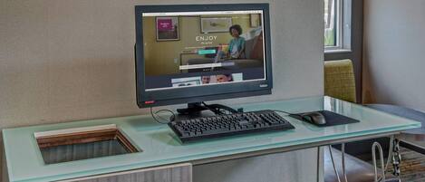 A modern hotel room with a glass desk, a desktop computer, a keyboard, and a mouse.