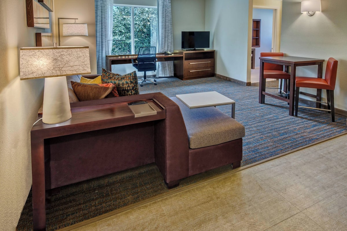 A hotel room with a sofa, a desk with a chair, a TV, and a dining table with chairs.