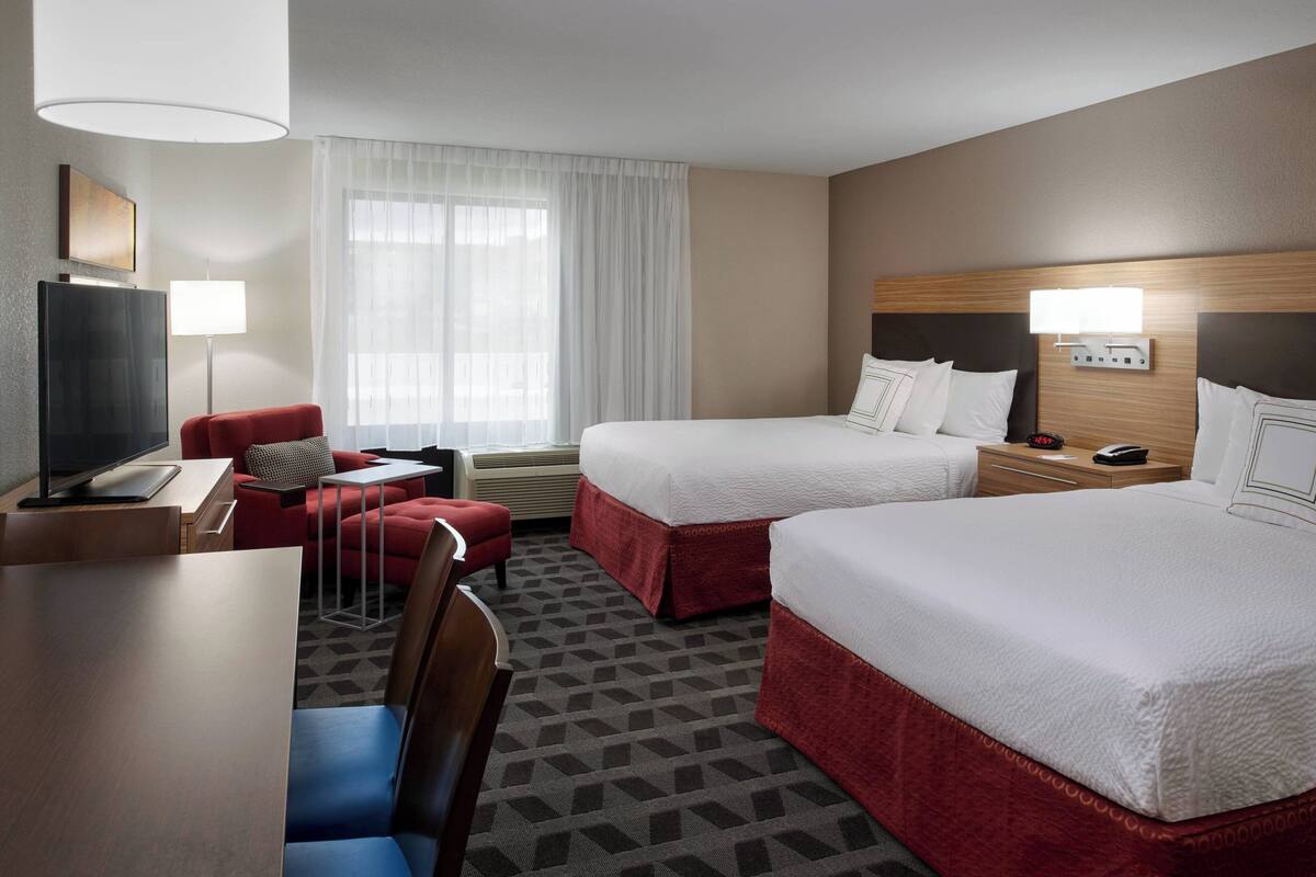 A hotel room with two beds, a desk, a TV, a chair, and a window with curtains.