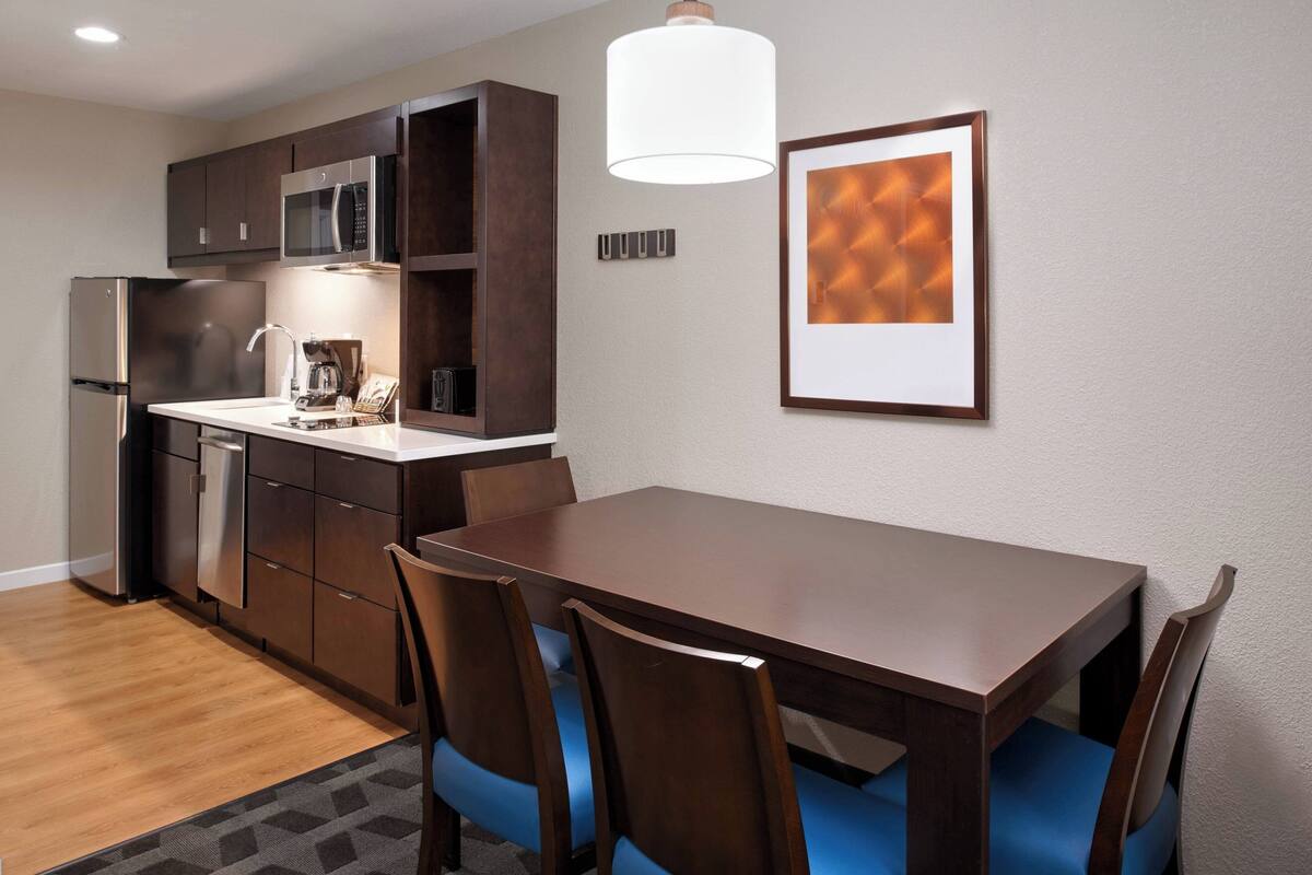 A modern kitchen with dark wooden cabinets, a stainless steel refrigerator, and a microwave. There is a dining table with chairs and a pendant light above it.