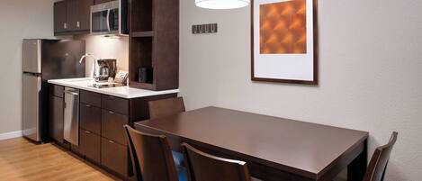 A modern kitchen with dark wooden cabinets, a stainless steel refrigerator, and a microwave. There is a dining table with chairs and a pendant light above it.