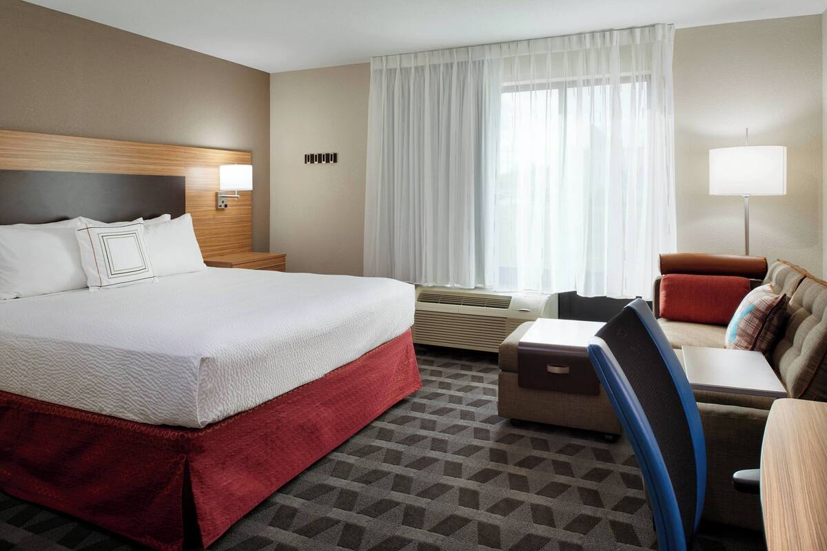 A hotel room with a large bed, a desk with a chair, a sofa, and a window with curtains.