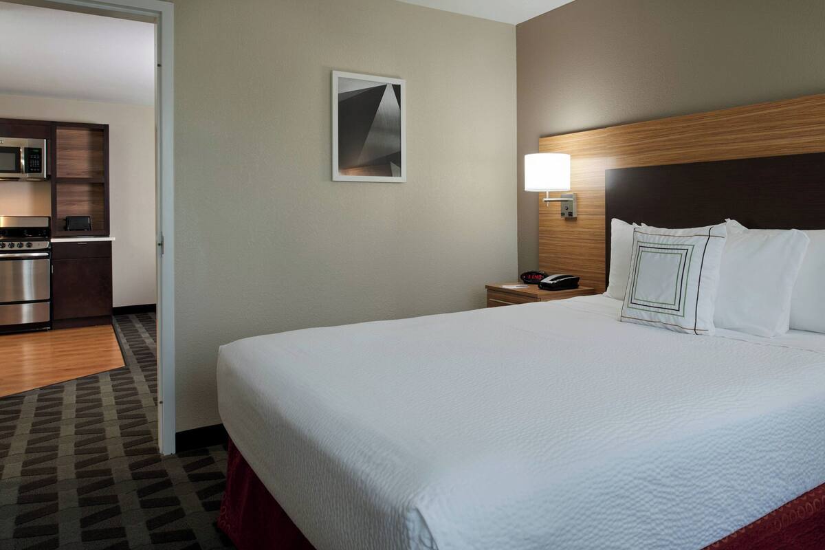 A hotel room with a large bed, a wooden headboard, a bedside table with a lamp, a microwave, and a small kitchenette.