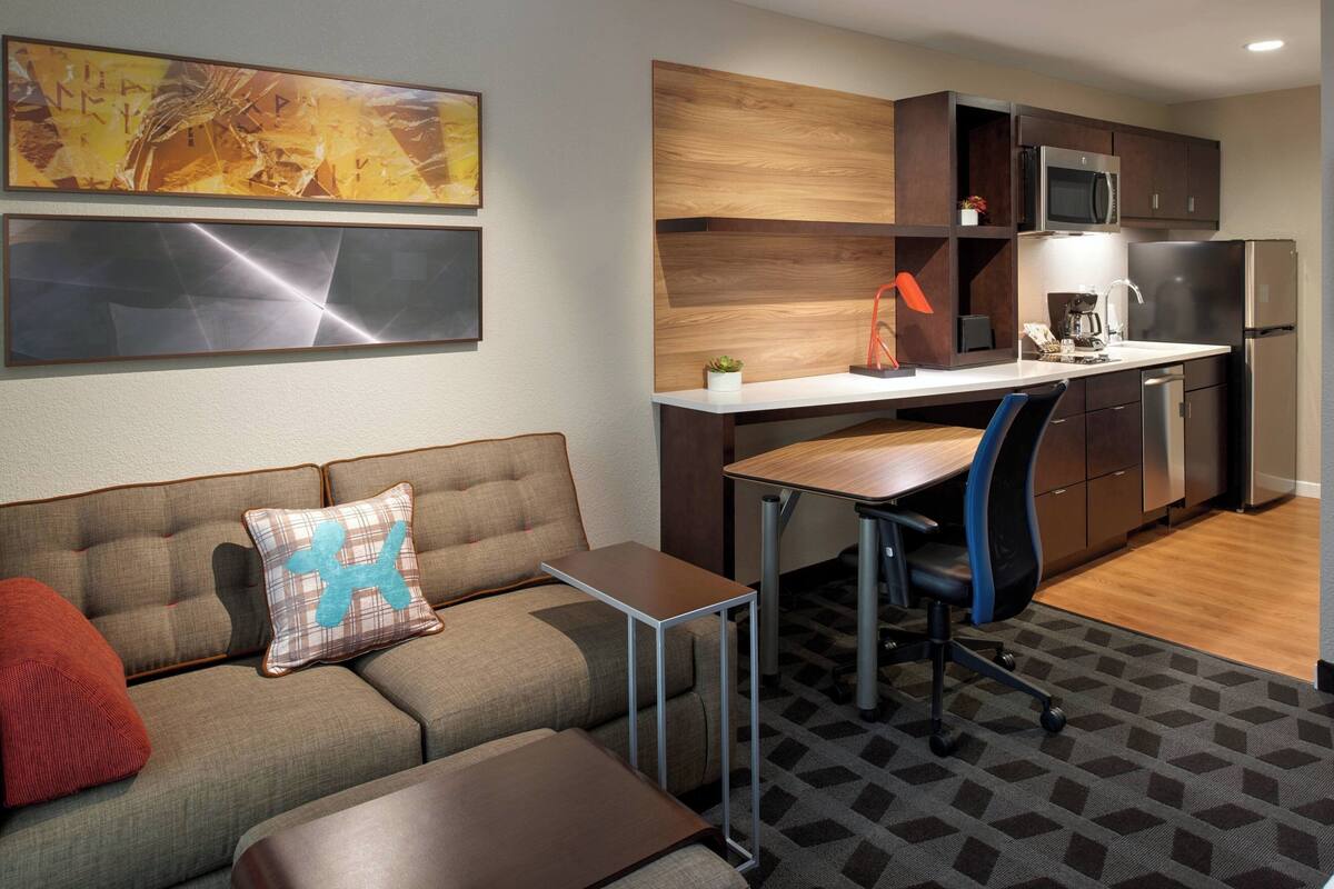 A modern kitchen with a microwave, refrigerator, and coffee maker. There is a sofa with a red pillow and a blue pillow with a white design. A small table is next to the sofa.
