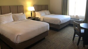 Quarto de hotel com duas camas, uma mesa de cabeceira com abajur, uma cadeira e uma janela com cortinas.