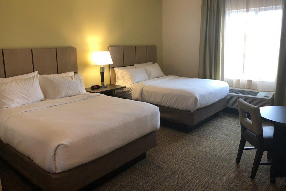 A hotel room with two beds, a bedside table with a lamp, a chair, and a window with curtains.