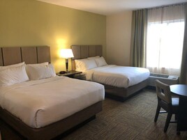 Quarto de hotel com duas camas, uma mesa de cabeceira com abajur, uma cadeira e uma janela com cortinas.