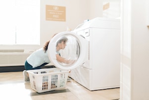 A person loading a white laundry basket into a front-loading washing machine in a room with a sign indicating free washer and dryer use.