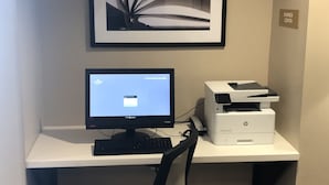 Uma área de trabalho com computador, impressora e uma obra de arte emoldurada em uma escrivaninha.