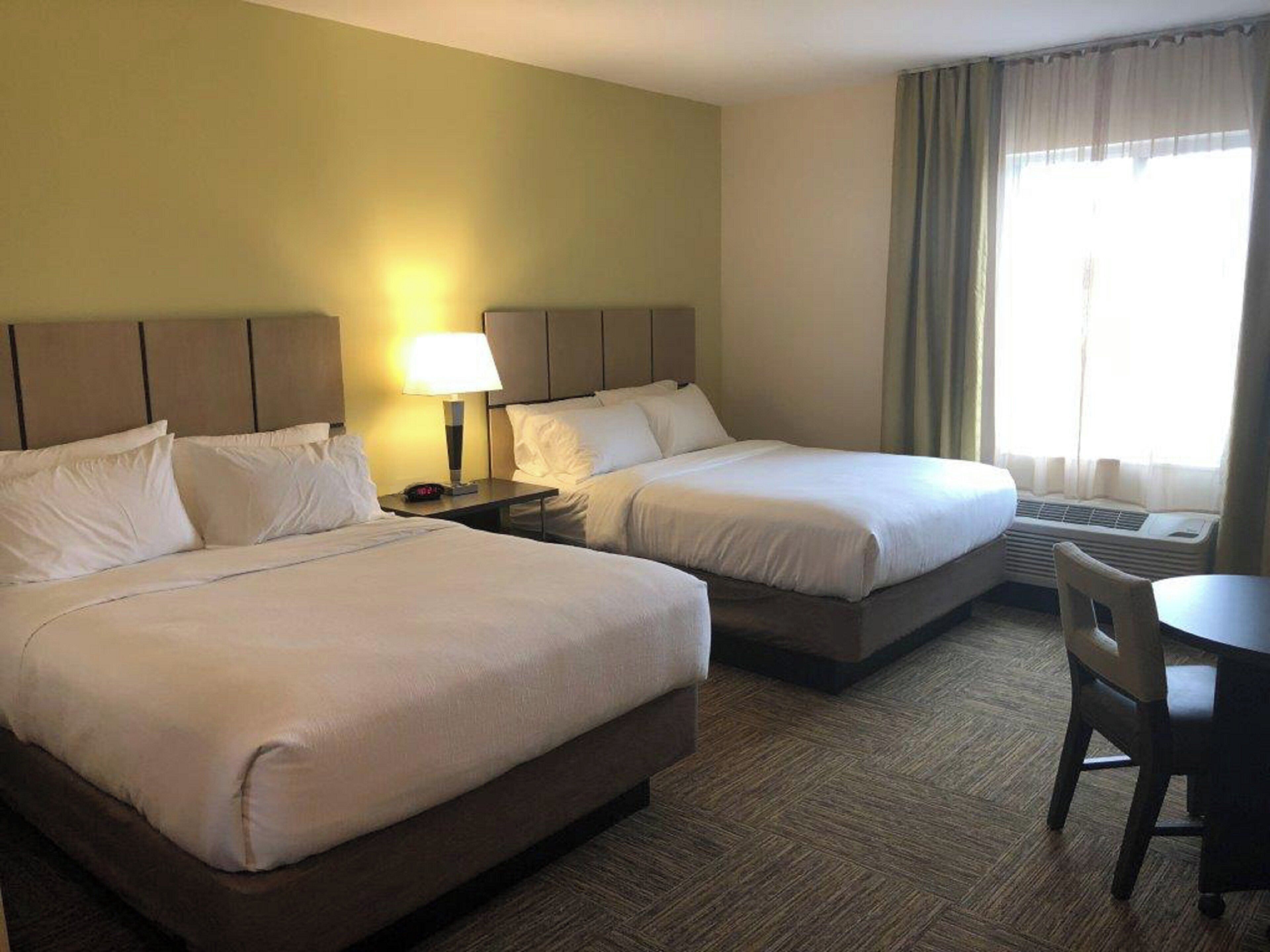 A hotel room with two beds, a bedside table with a lamp, a chair, and a window with curtains.