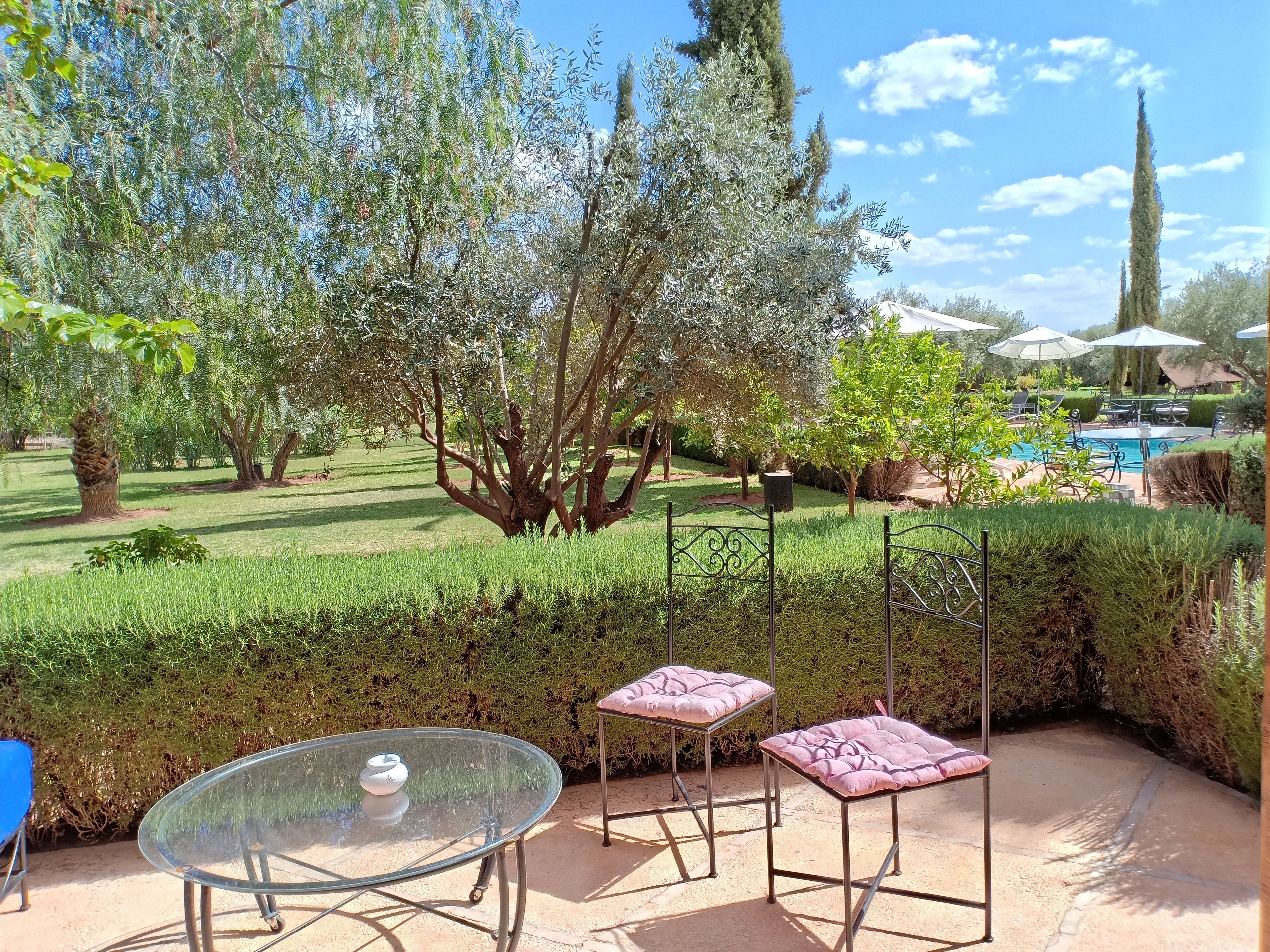 An outdoor patio with a glass table and two cushioned chairs, surrounded by well-maintained hedges and trees.