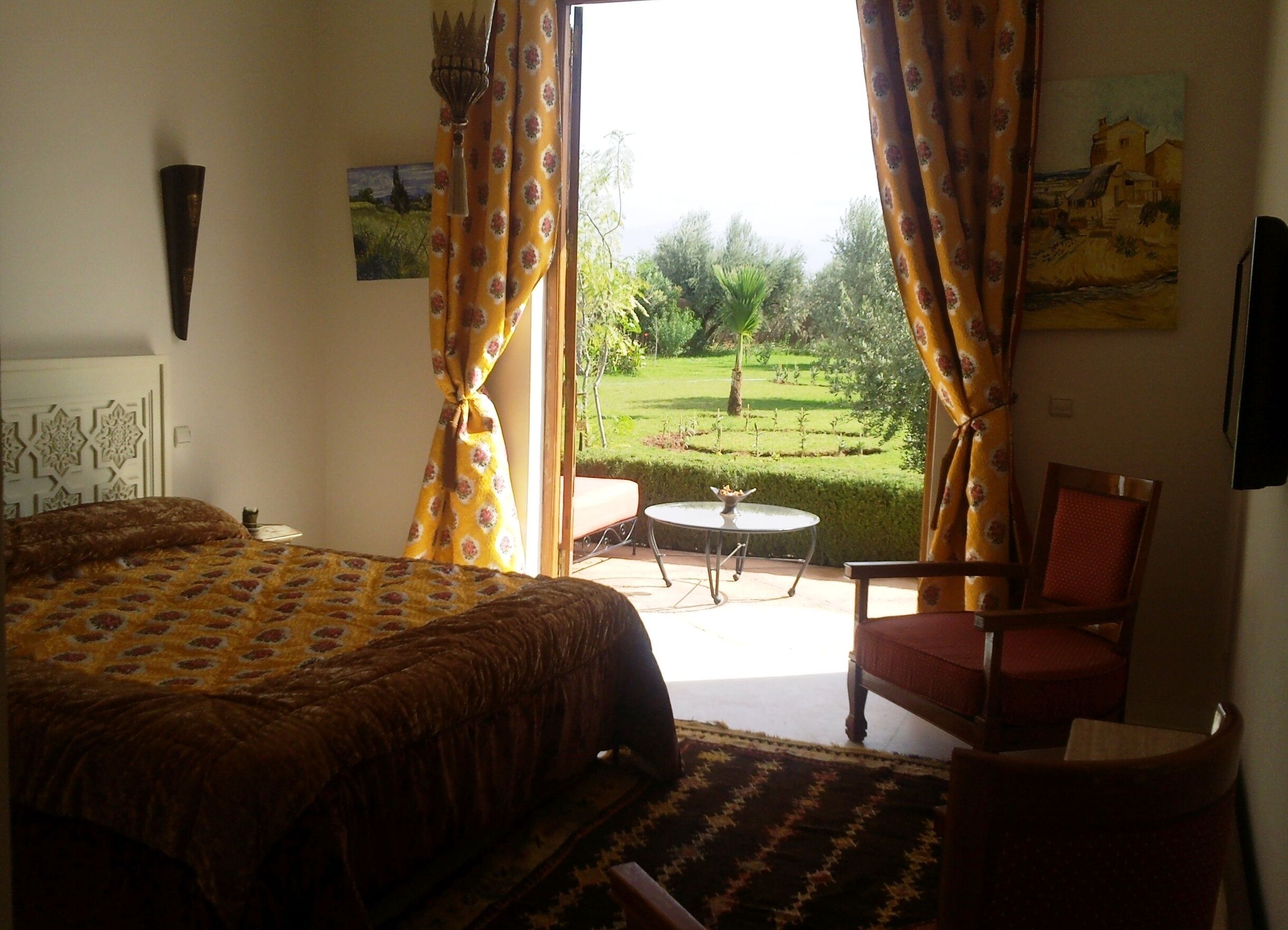 A bedroom with a bed, a chair, a small table, and a view of a garden through open curtains.