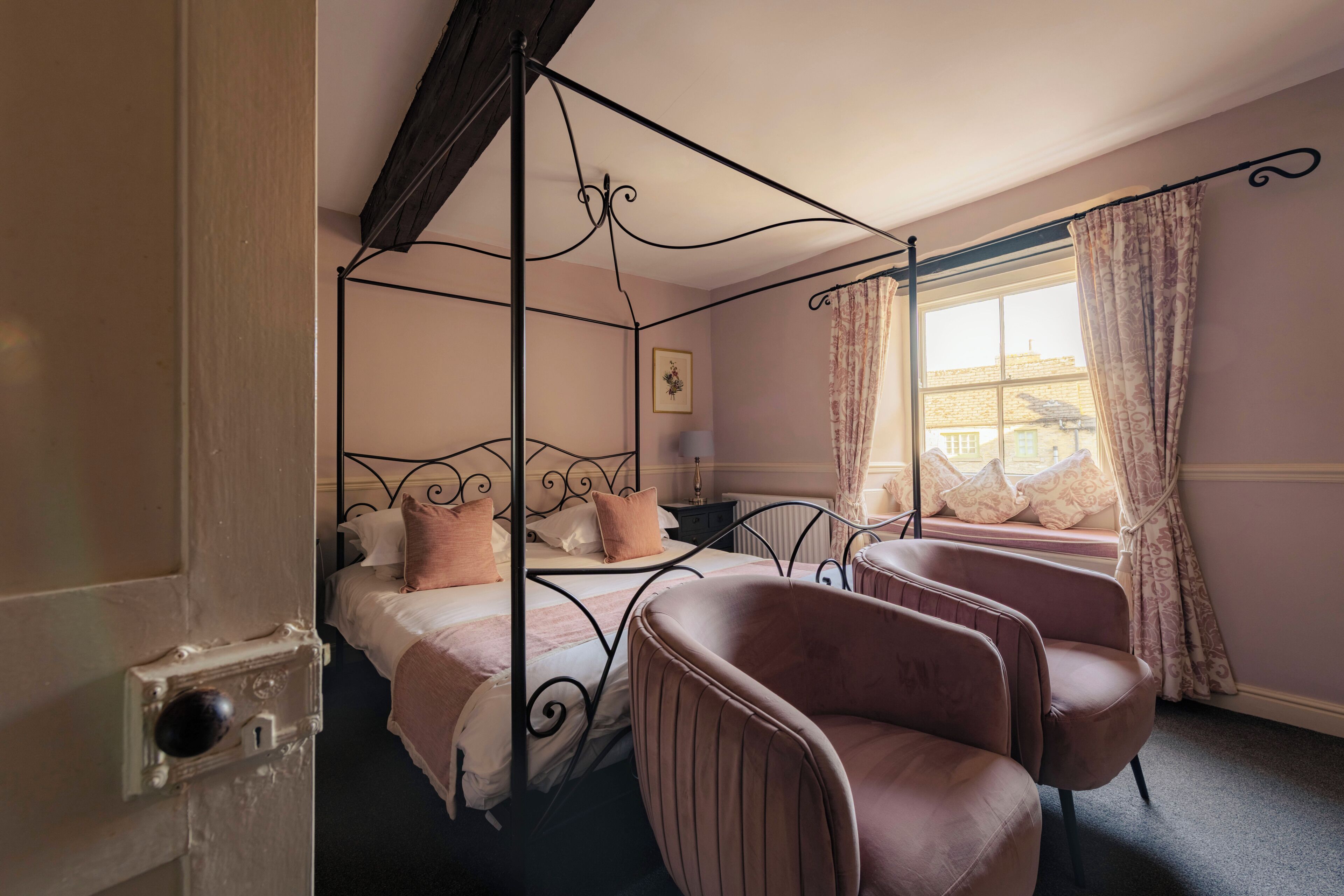 A four-poster bed with a headboard, a sitting area with two chairs, a window with curtains, and a door with a lock.