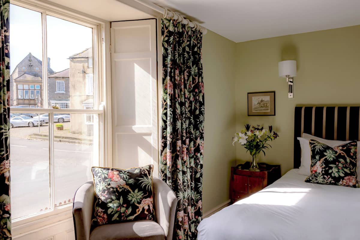 A hotel room with a bed, a chair with floral cushions, a nightstand with a vase of flowers, and a window with curtains.