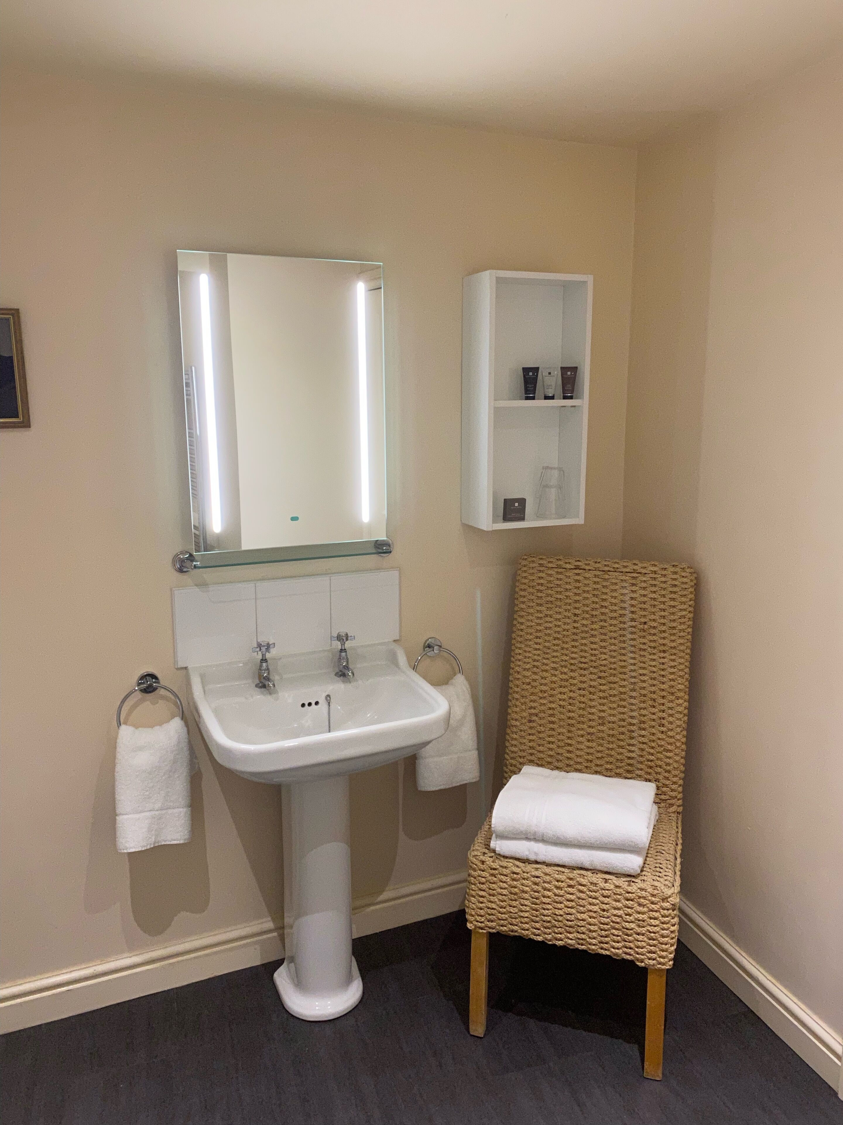 A bathroom with a pedestal sink, a mirror, a towel rack, a wicker chair, and a small shelf with toiletries.