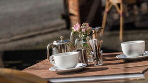Una mesa dispuesta con tetera, tazas y un florero con flores, rodeada de sillas de mimbre.