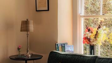 A cozy room with a sofa, a small table with a lamp, a framed picture, and a window with a view of trees.