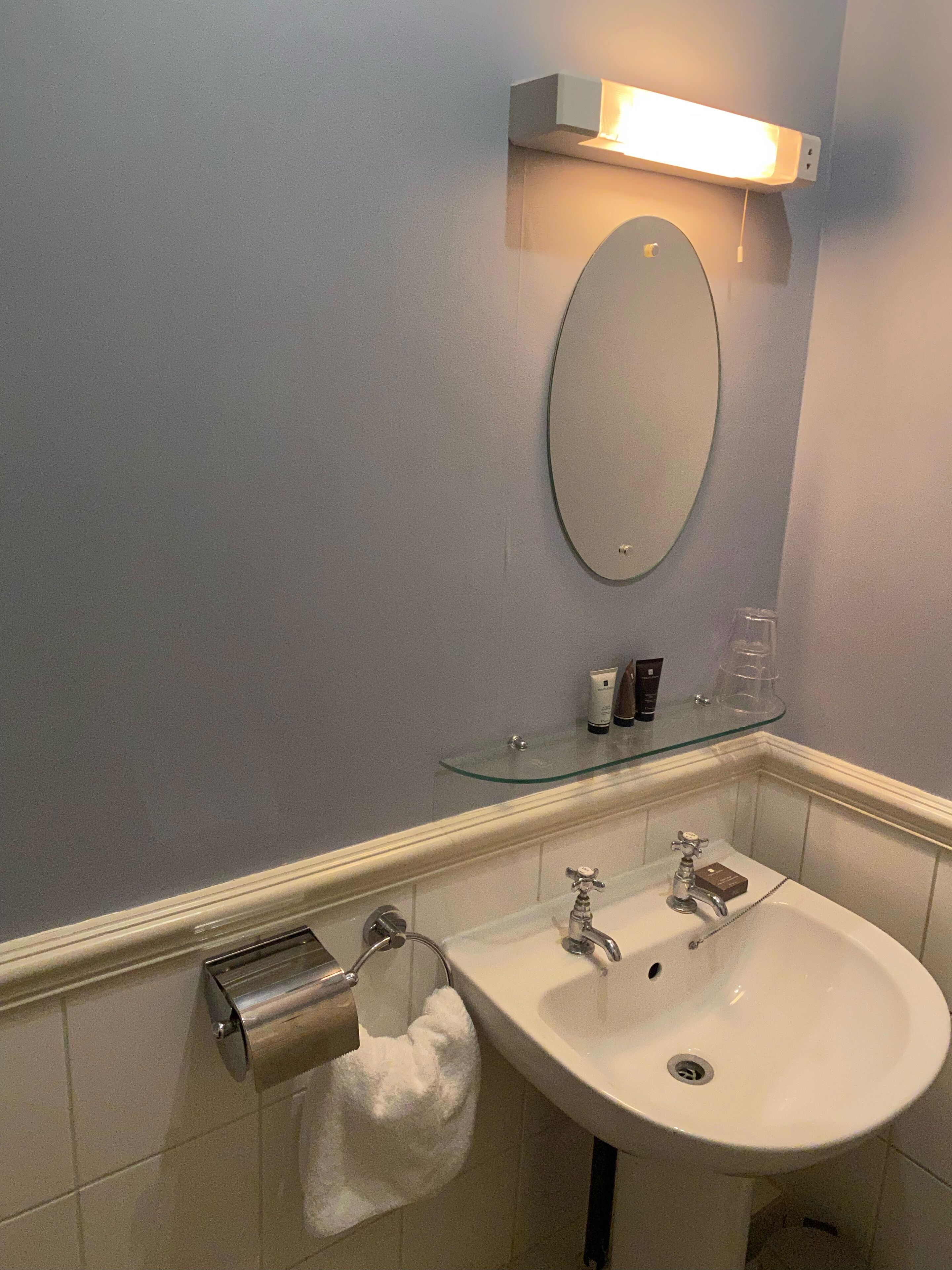 Un baño con un lavabo blanco, dos grifos, un espejo y un estante de vidrio con artículos de aseo.