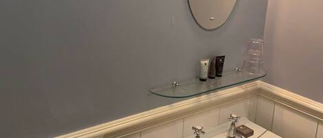 A bathroom with a white sink, two faucets, a mirror, and a glass shelf with toiletries.