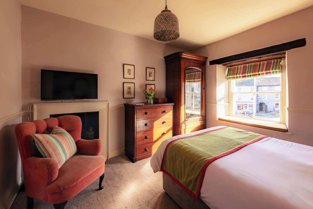 A bedroom with a bed, a chair, a dresser, a television, and a window with a view of the street.