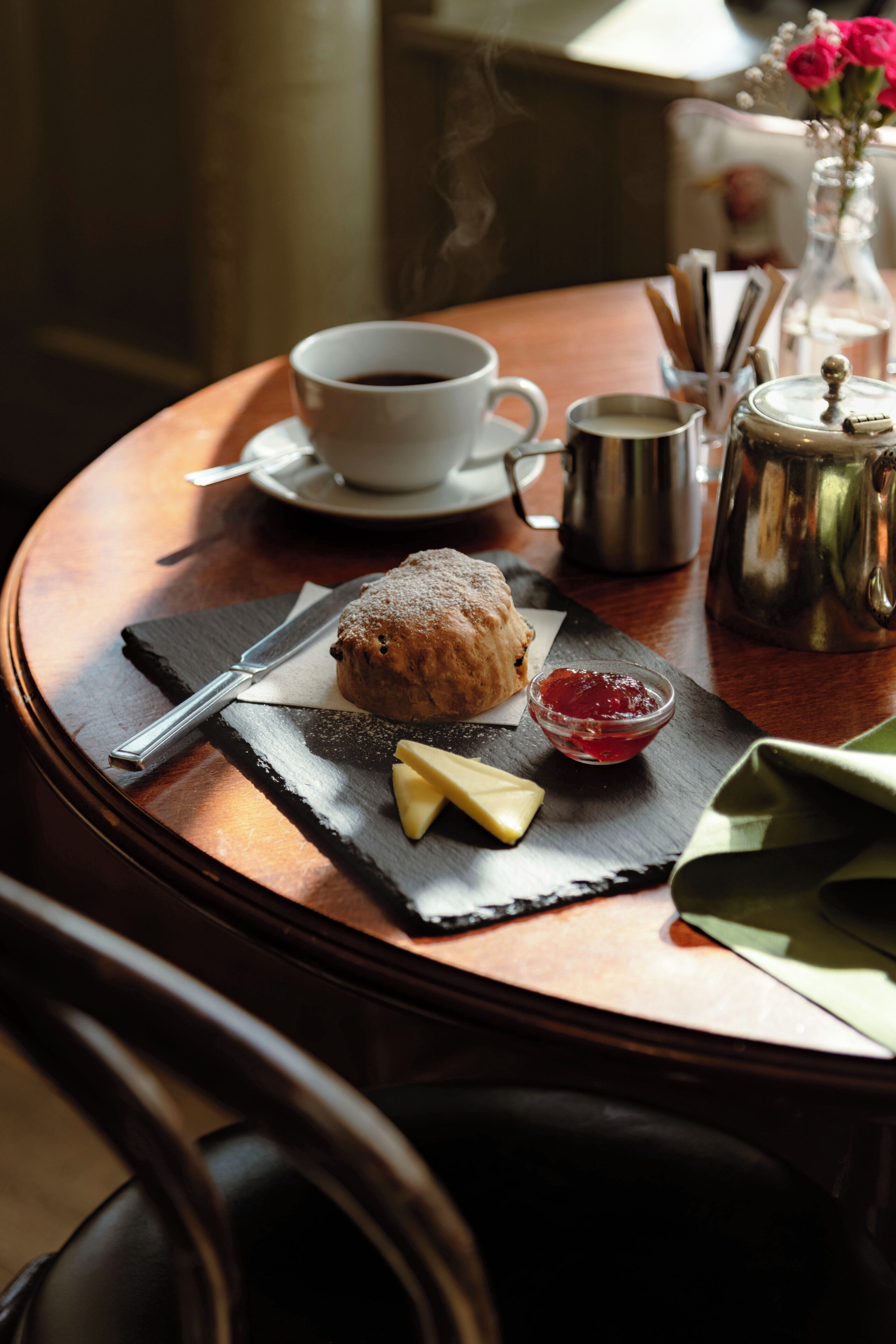 A round table with a pastry, a cup of coffee, a teapot, and a small vase with flowers.