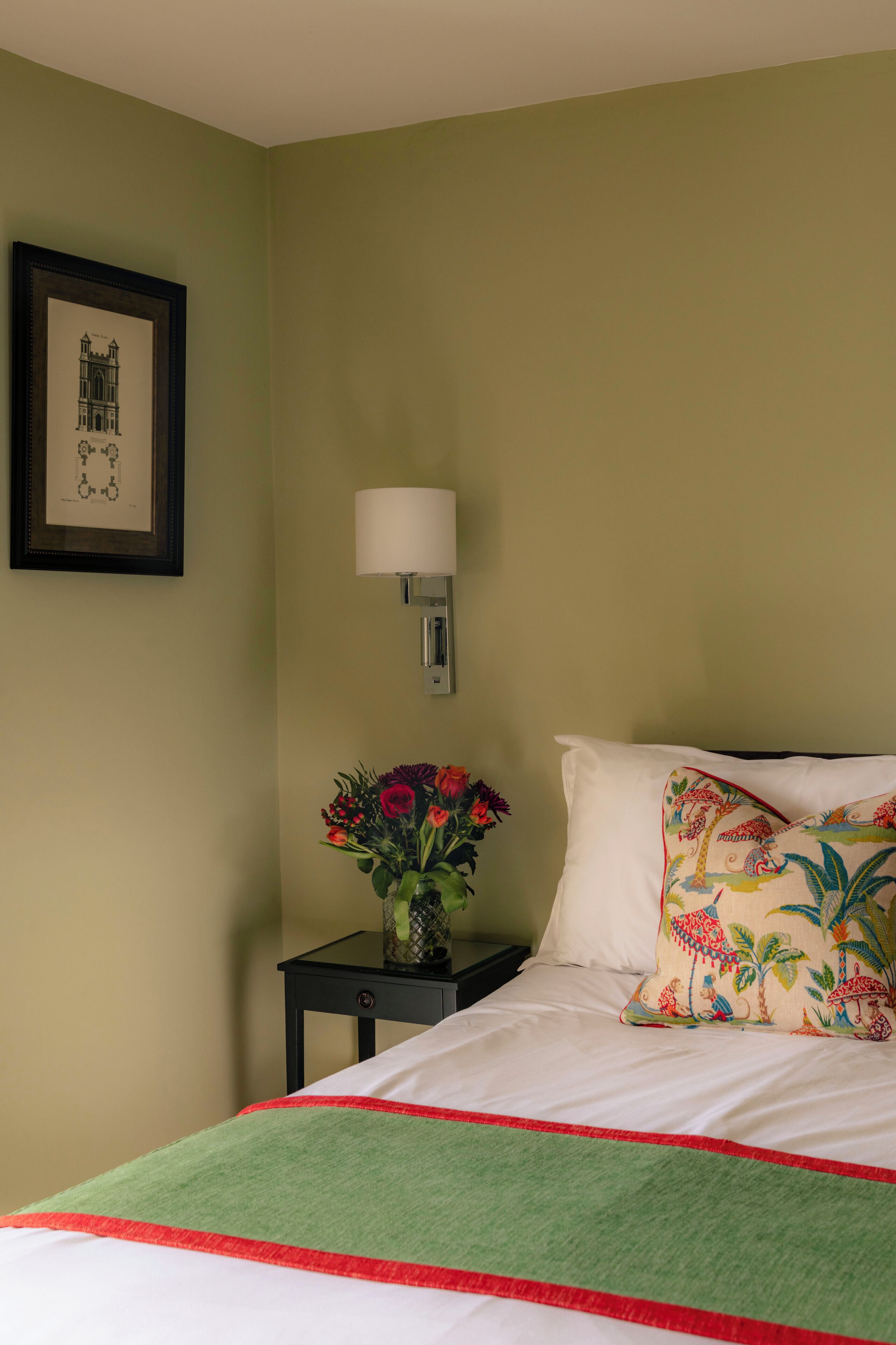 A neatly made bed with a green and red bedspread, a floral pillow, a vase with flowers on a bedside table, and a framed picture on the wall.