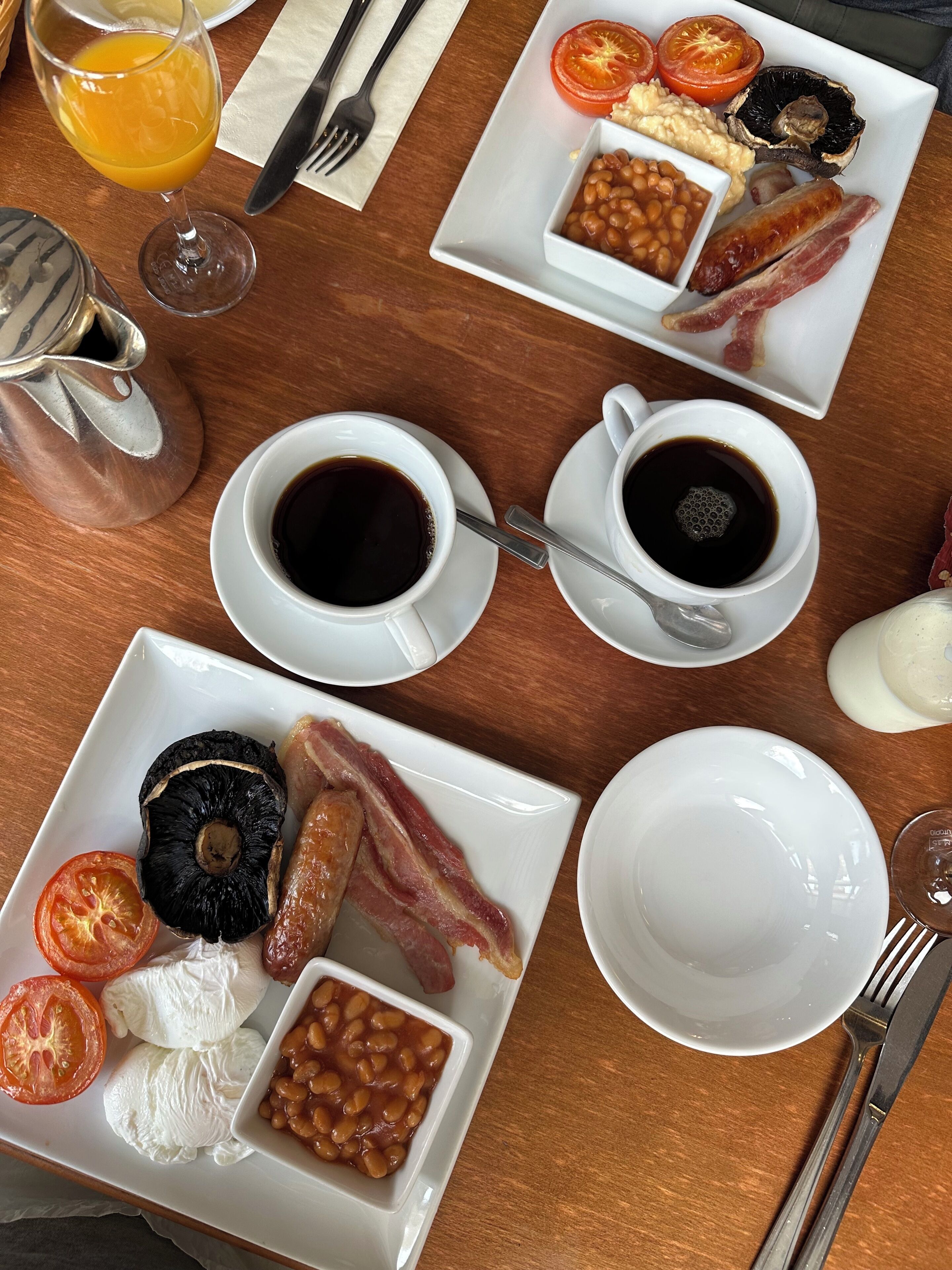 A breakfast setting with plates of eggs, bacon, tomatoes, beans, and mushrooms, along with cups of coffee and a glass of orange juice.