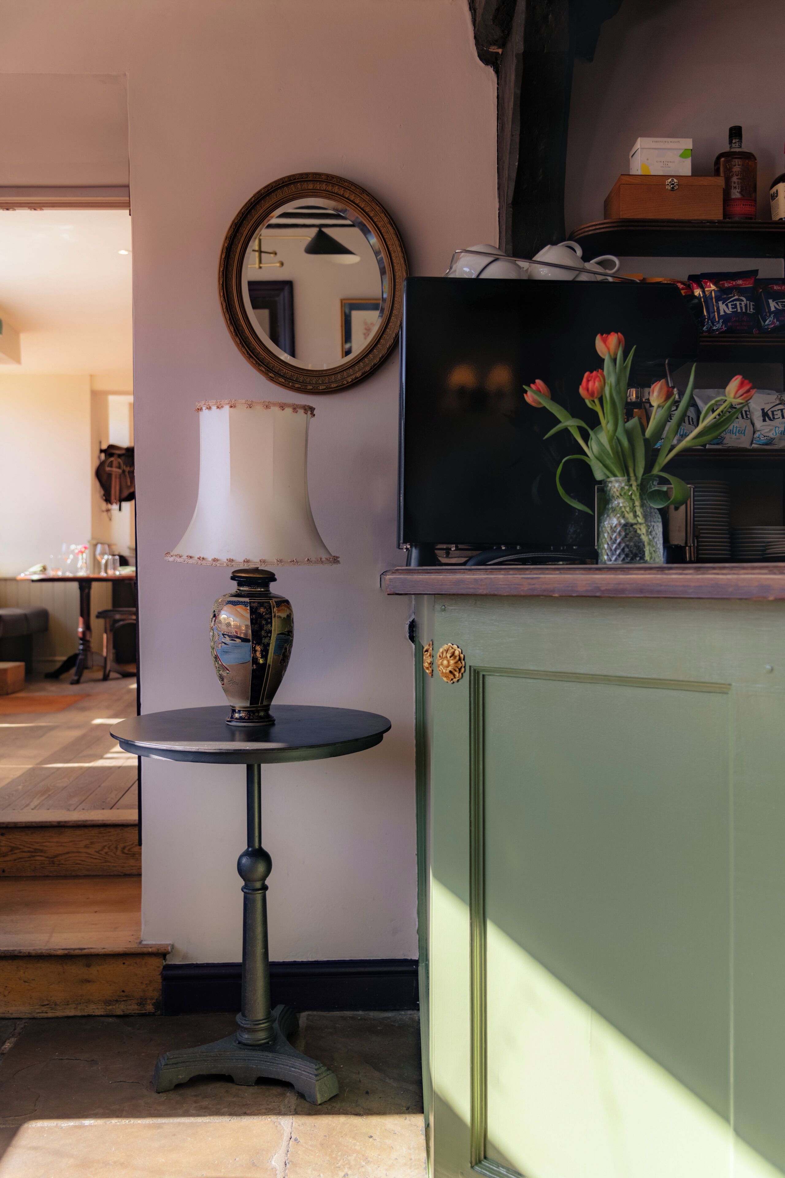 Une pièce avec un meuble vert, un miroir rond, une table avec une lampe et un vase rempli de fleurs.