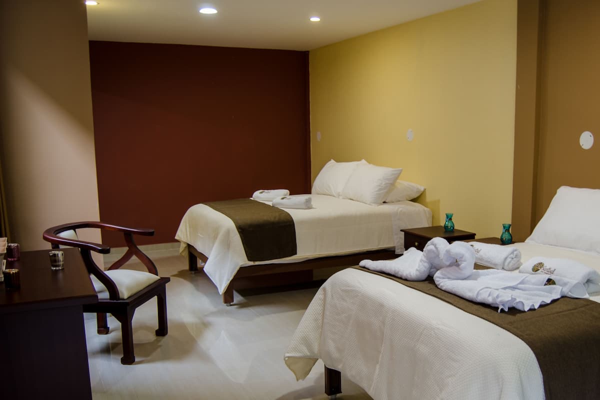 Quarto de hotel com duas camas, uma cadeira de madeira, uma mesinha e uma mesa de cabeceira com dois vasos azul-turquesa.