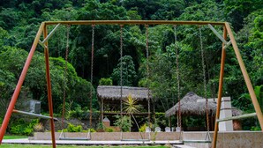 A swing set in a lush garden with a thatched-roof structure in the background. - Santval (Cumanda)