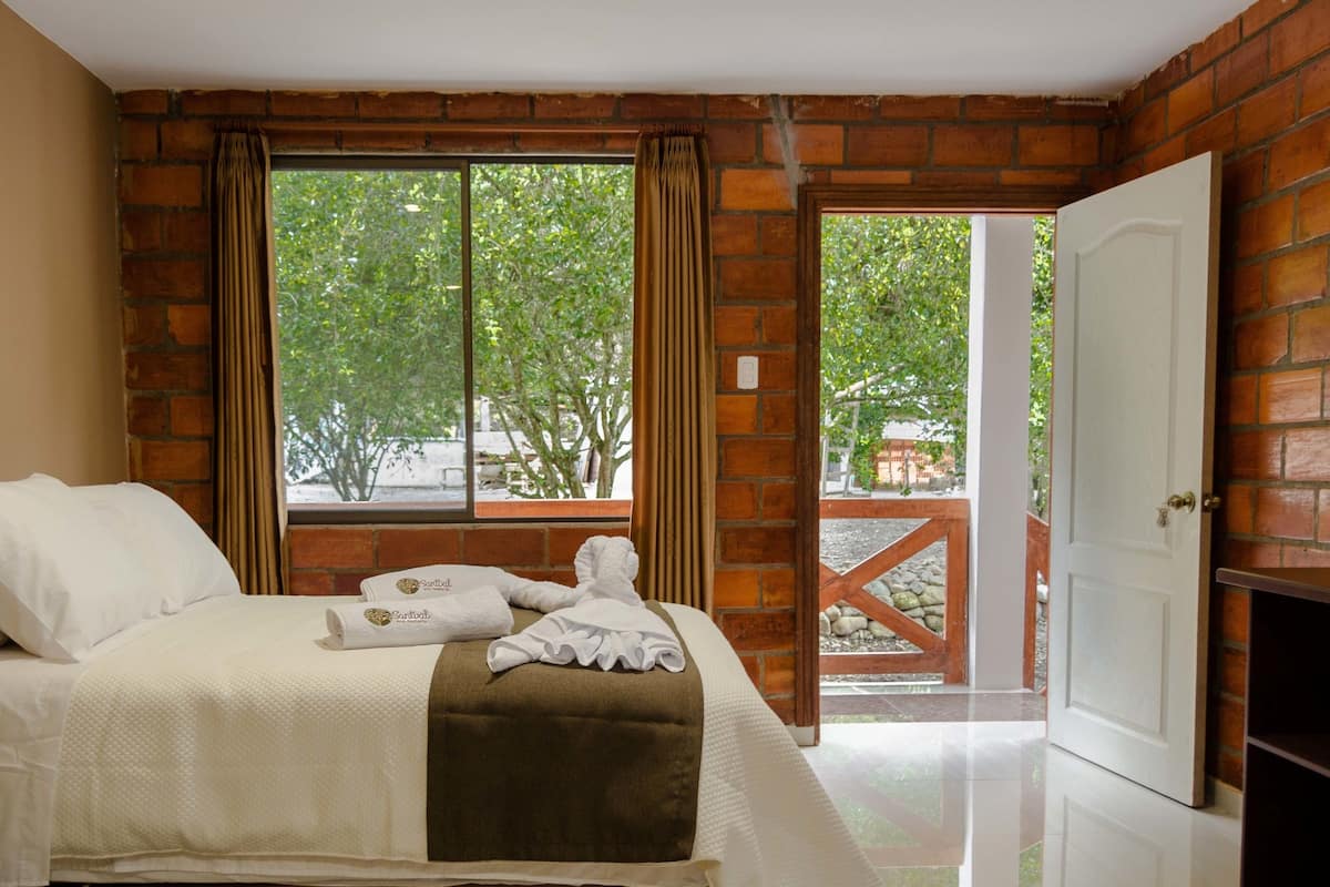 Quarto de hotel com cama, janela com cortinas, porta e vista para o exterior.