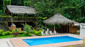 A swimming pool with a thatched-roof structure and a brick building in a lush garden setting. - Santval (Cumanda)